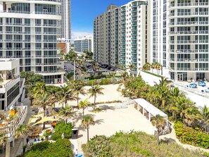 Un immeuble résidentiel de grande hauteur doté de nombreux balcons, une plage de sable bordée de palmiers et un parking.