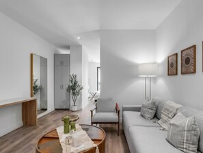 Ett modernt vardagsrum med en grå soffa, ett träbord och en lampa. Det finns en grå fåtölj och ett litet bord med dekorativa föremål. Rummet har vita väggar och ett trägolv.
