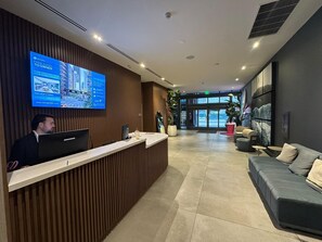 En hotellobby med en digital skärm som visar information om hotellet, en receptionsdisk, en sittgrupp med en soffa och fåtöljer samt ett stort fönster med utsikt över gatan.
