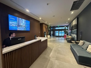 Un espace d’accueil d’hôtel doté d’un écran numérique affichant des informations sur l’établissement, d’un comptoir de réception, d’un coin salon avec un canapé et des fauteuils, et d’une grande fenêtre donnant sur la rue.
