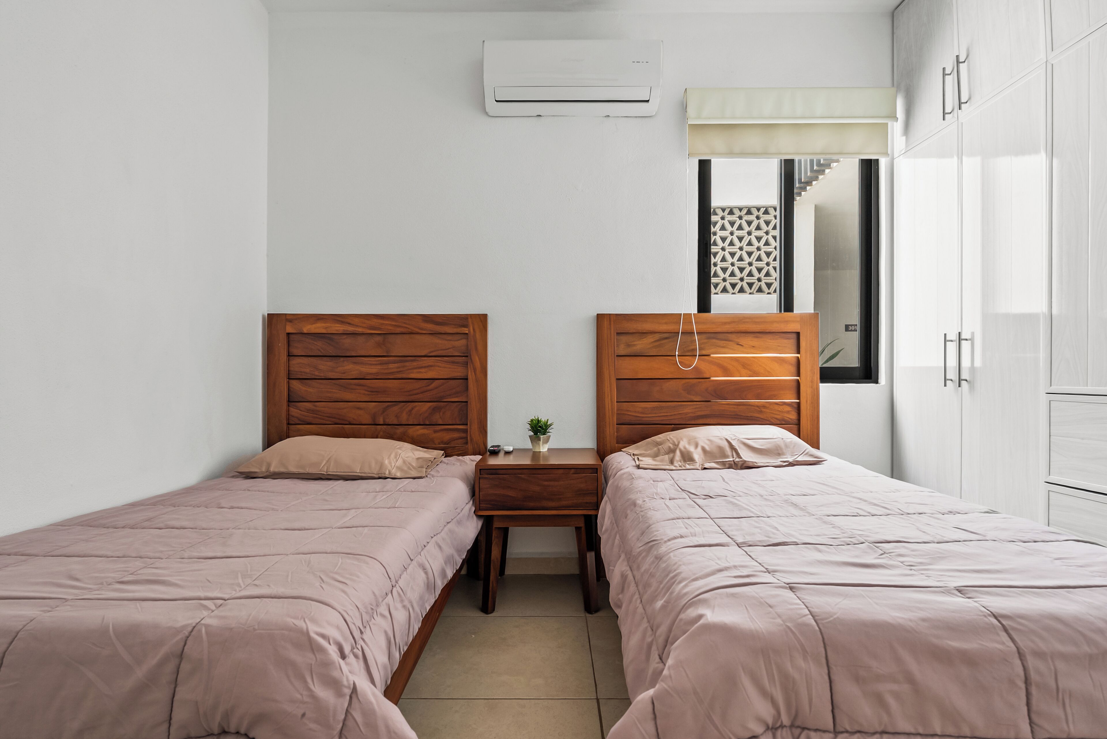 A bedroom with two wooden beds, a nightstand, a small table with a plant, and a window with a geometric pattern.