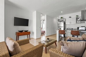 Ein modernes Wohnzimmer mit Flachbildfernseher, Korbmöbeln, einem Glastisch und einer Küchenzeile mit Barhocker.