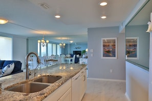 A modern kitchen with granite countertops, stainless steel sinks, and a dining area with a white table and chairs. - Serene Saltwater Sanctuary | Inlet #607 (New Smyrna Beach)