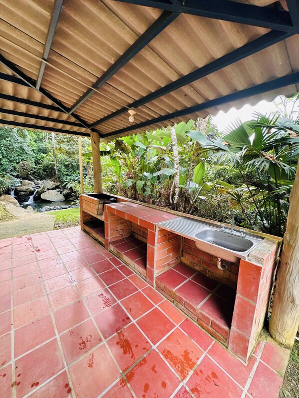A covered outdoor area with a brick-built sink and bench, surrounded by lush greenery and a small waterfall. - Creekvista Vista y Encanto del Charco (Cocorná)