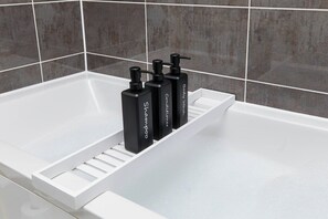 A bathroom with a white bathtub, a white shelf, and three black bottles of shampoo. - Luxury 2 Bed Apt at Victoria Riverside | Parking (Leeds)