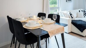 Un comedor con una mesa dispuesta para cuatro, sillas y, al fondo, se ve un sofá blanco y una ventana.