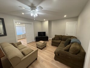 Un salon Ă©quipĂ© dâun ventilateur de plafond, dâun tĂ©lĂ©viseur, de deux canapĂ©s et dâune porte donnant sur une autre piĂšce.