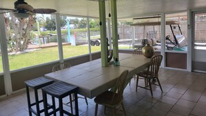 Eine überdachte Terrasse mit Tisch und Stühlen, einem Deckenventilator und Blick auf einen Garten mit Golfwagen und Zaun.