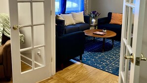 Puerta abierta que da a una sala de estar con un sofá de color oscuro, una mesa redonda de centro y un tapete azul estampado.
