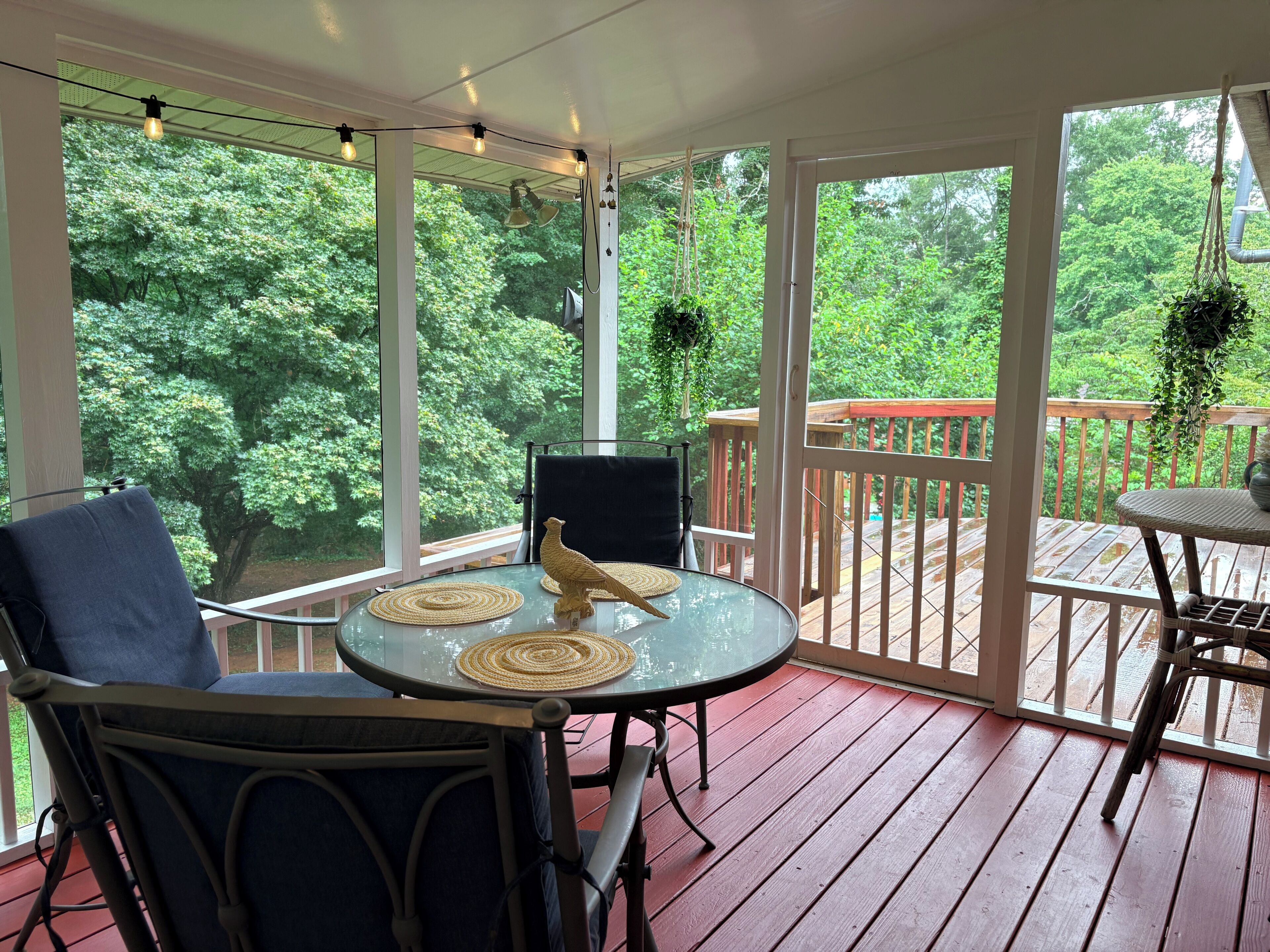 A covered outdoor seating area with a table and chairs, overlooking a wooden deck and lush greenery.