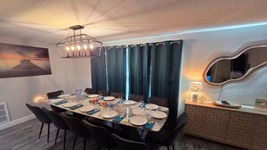Uma sala de jantar com uma mesa comprida posta para seis, cortinas escuras, um lustre moderno e um espelho fixado na parede.