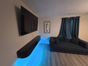 Ein modernes Wohnzimmer mit einem wandmontierten Flachbildfernseher, einem dunklen Sofa und einer gerahmten Meerpferd-Kunst.'