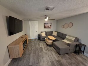 Ein modernes Wohnzimmer mit Ecksofa, Fernseher, Deckenventilator und einer Tür.