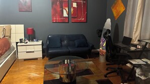 A room with a bed, a sofa, a glass coffee table, a desk with a chair, and artwork on the walls.