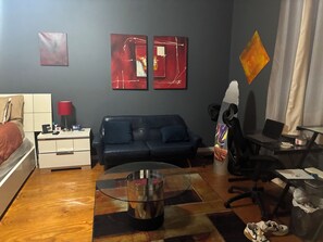 A room with a bed, a sofa, a glass coffee table, a desk with a chair, and artwork on the walls.