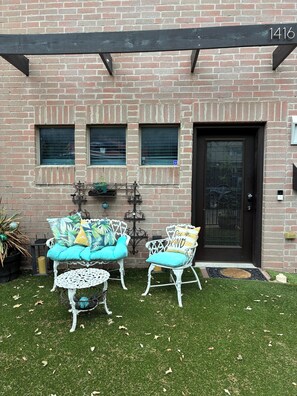 A brick building with a black door, two white wicker chairs with teal cushions, and a small table.