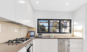 A modern kitchen with white cabinets, a stainless steel dishwasher, a gas stove, and a window with a view of buildings and trees. - MetaWiseBnB 2BR Penthouse style near Bondi Beach & Westfield (Bondi Junction)