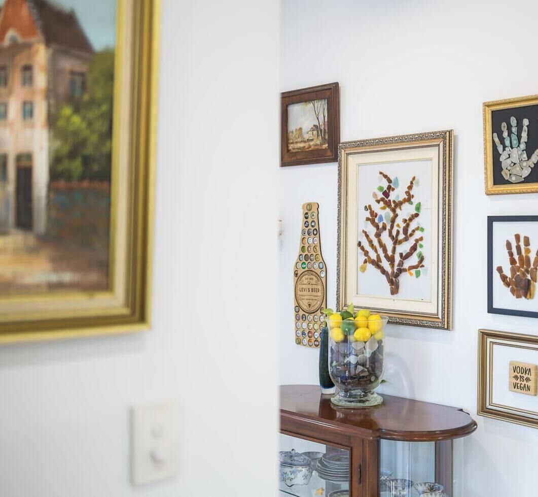 Un mur orné de divers tableaux encadrés, une décoration en forme de bouteille et un vase rempli de fleurs jaunes.