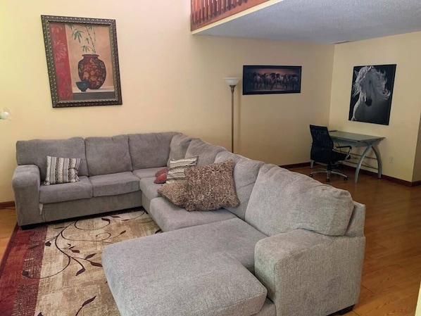 A living room with a grey sectional sofa, a small table, and a chair. There are framed pictures on the walls.