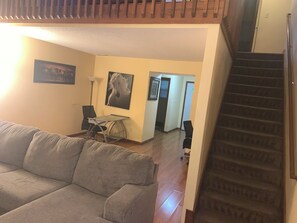 Ein Wohnzimmer mit einer Couch, einem Schreibtisch und einem Sessel, einer Treppe und einem Flur, der in einen anderen Raum führt.