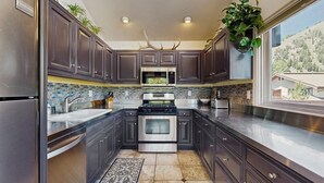 A modern kitchen with dark cabinets, stainless steel appliances, and a view of a mountainous landscape. - Spacious home near town w/ private hot tub- next to trails & river (Ketchum)