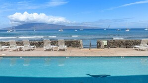 Ein Pool mit Liegestühlen, einer Steinmauer und Segelbooten auf dem Wasser.