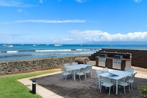 Eine Terrasse mit weißen Stühlen und einem Tisch, mit Blick auf das Meer und Boote in der Ferne.