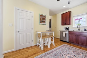 A kitchen with wooden cabinets, a white door, a small table with stools, a coffee maker, and a rug.