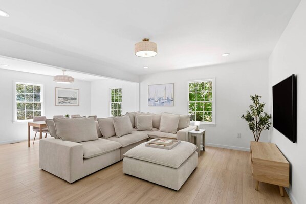 A spacious living room with a sectional sofa, a dining area with a table and chairs, large windows with greenery outside, and a mounted television on the wall.