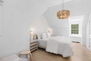 Ein modernes Schlafzimmer mit schrägem Dach, einem Bett mit weißer Bettwäsche, Nachttischlampen, einem kleinen hölzernen Hocker und einem großen Fenster mit Blick ins Grüne.