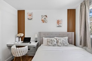 A bedroom with a bed, a desk with a laptop, a chair, and artwork on the wall.