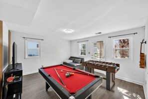 A pool table, foosball table, and a sofa in a room with white walls and windows.