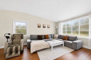 A living room with a massage chair, grey sofa, white coffee table, and large windows.