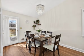 A dining room with a wooden table set for six, surrounded by chairs, a chandelier, and wall-mounted shelves with plants.