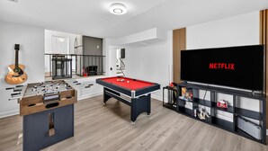 Una sala de estar moderna con mesa de fútbol, mesa de billar y una gran pantalla de televisión mostrando 'NETFLIX'.