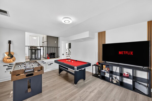 A modern living room with a foosball table, pool table, and a large TV screen displaying 'NETFLIX'.