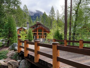 Eine hölzerne Brücke führt zu einer Lodge mit einem Steinkamin, umgeben von dichtem Wald und Bergen.