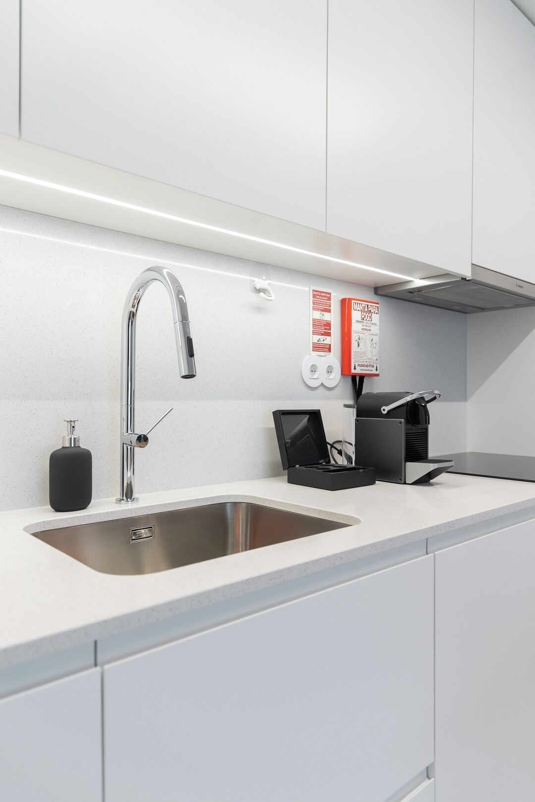 Eine moderne Küche mit Spüle aus Edelstahl, Wasserhahn, Seifenspender, Kaffeemaschine und einem Brandschutzplakat.
