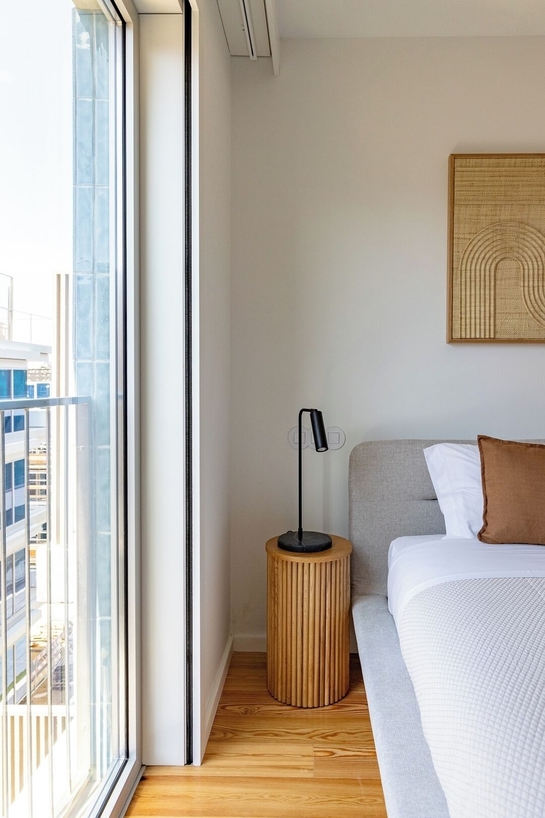 A modern bedroom with a large window, a round wooden nightstand, a black floor lamp, a grey sofa, and a white bed.
