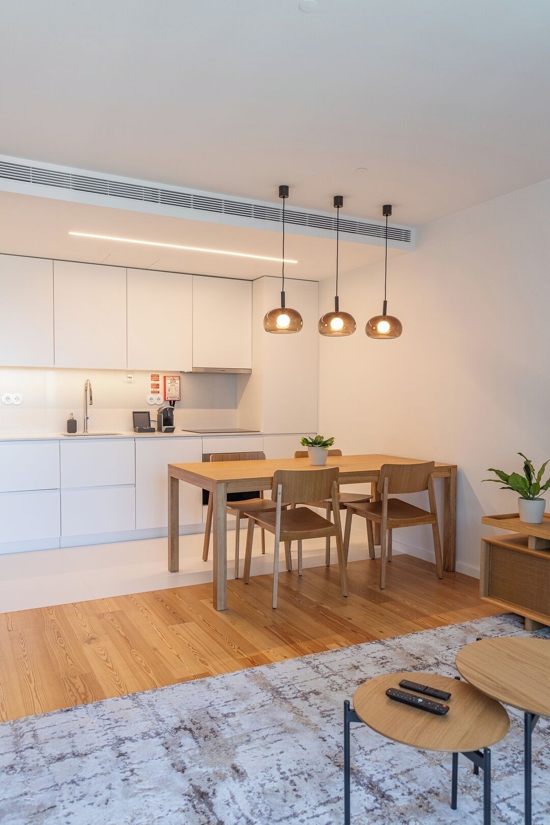 A modern kitchen with white cabinets, a wooden dining table, and pendant lights.