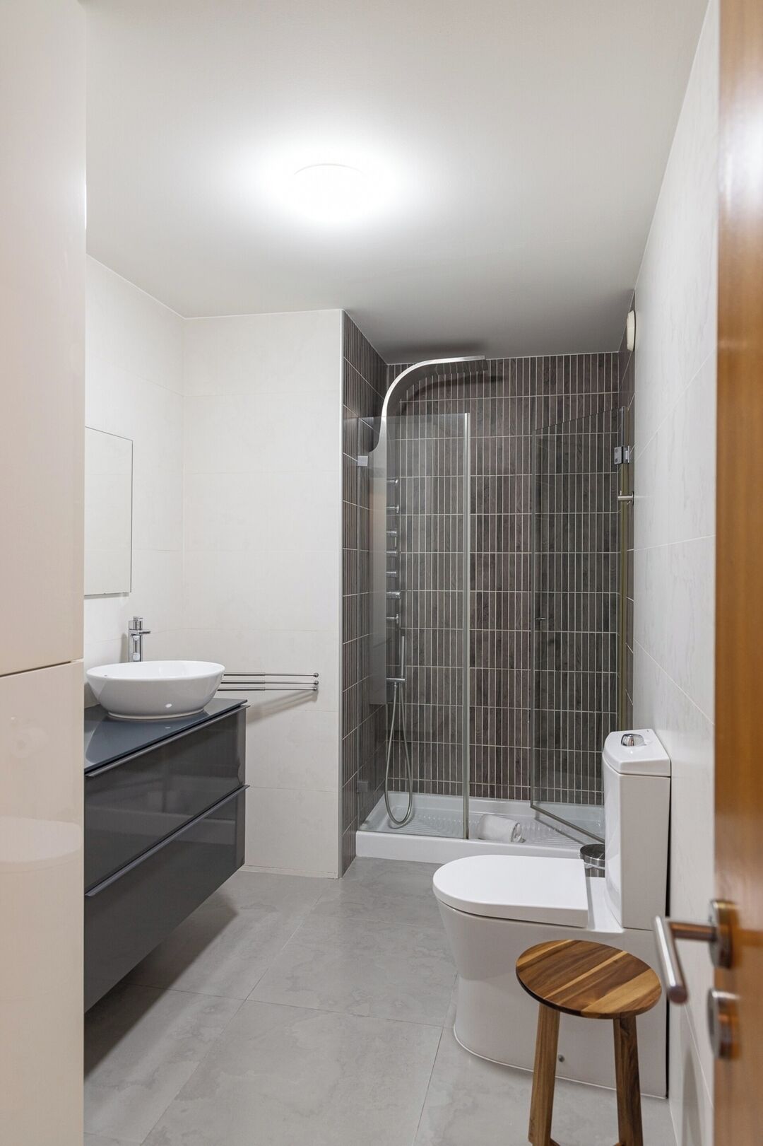 A modern bathroom with a white sink, a glass shower enclosure, and a wooden stool.