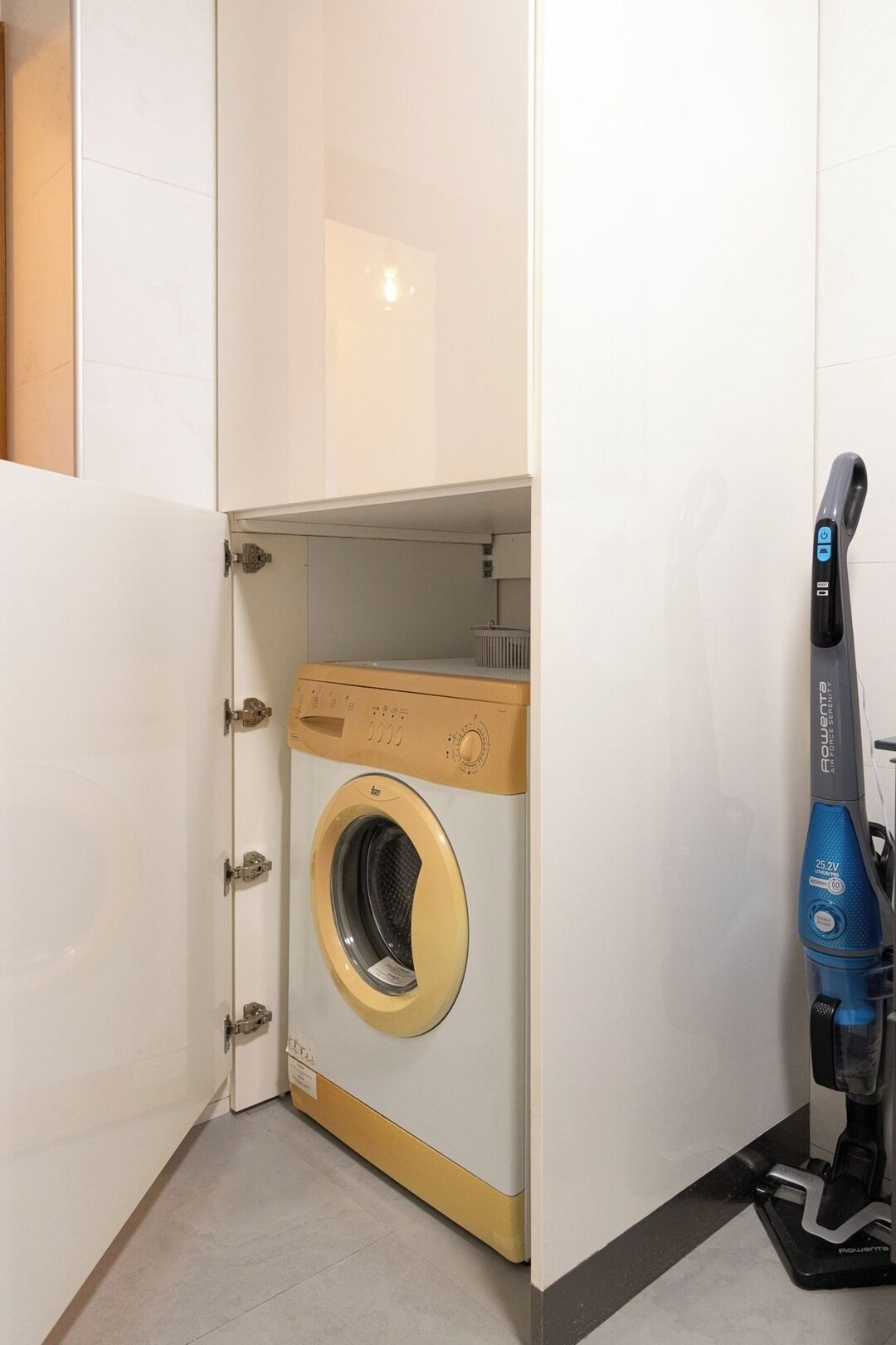 A compact, built-in washing machine with a control panel, a vacuum cleaner, and a small trash bin in a corner of a room with white walls and a tiled floor.