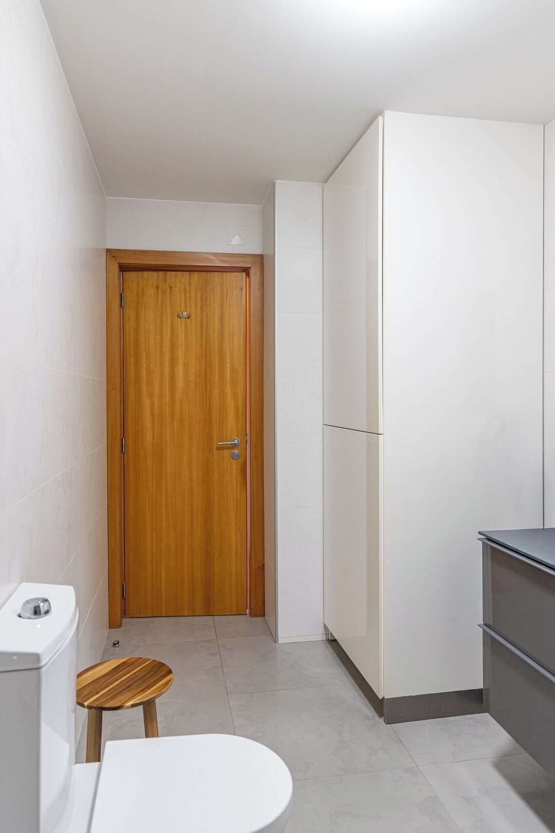 A narrow hallway with a wooden door, a white toilet, a small wooden stool, and a white cabinet.