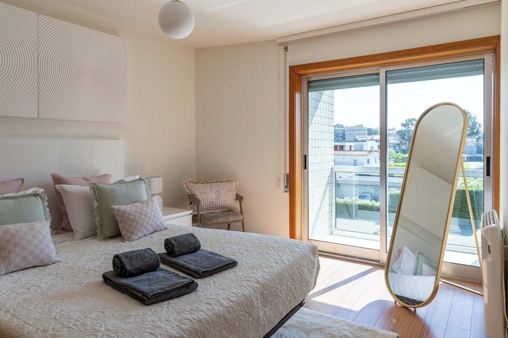 Ein Schlafzimmer mit Bett, Kissen, einem Sessel, einem großen Spiegel und Blick auf Gebäude durch eine Schiebetür.