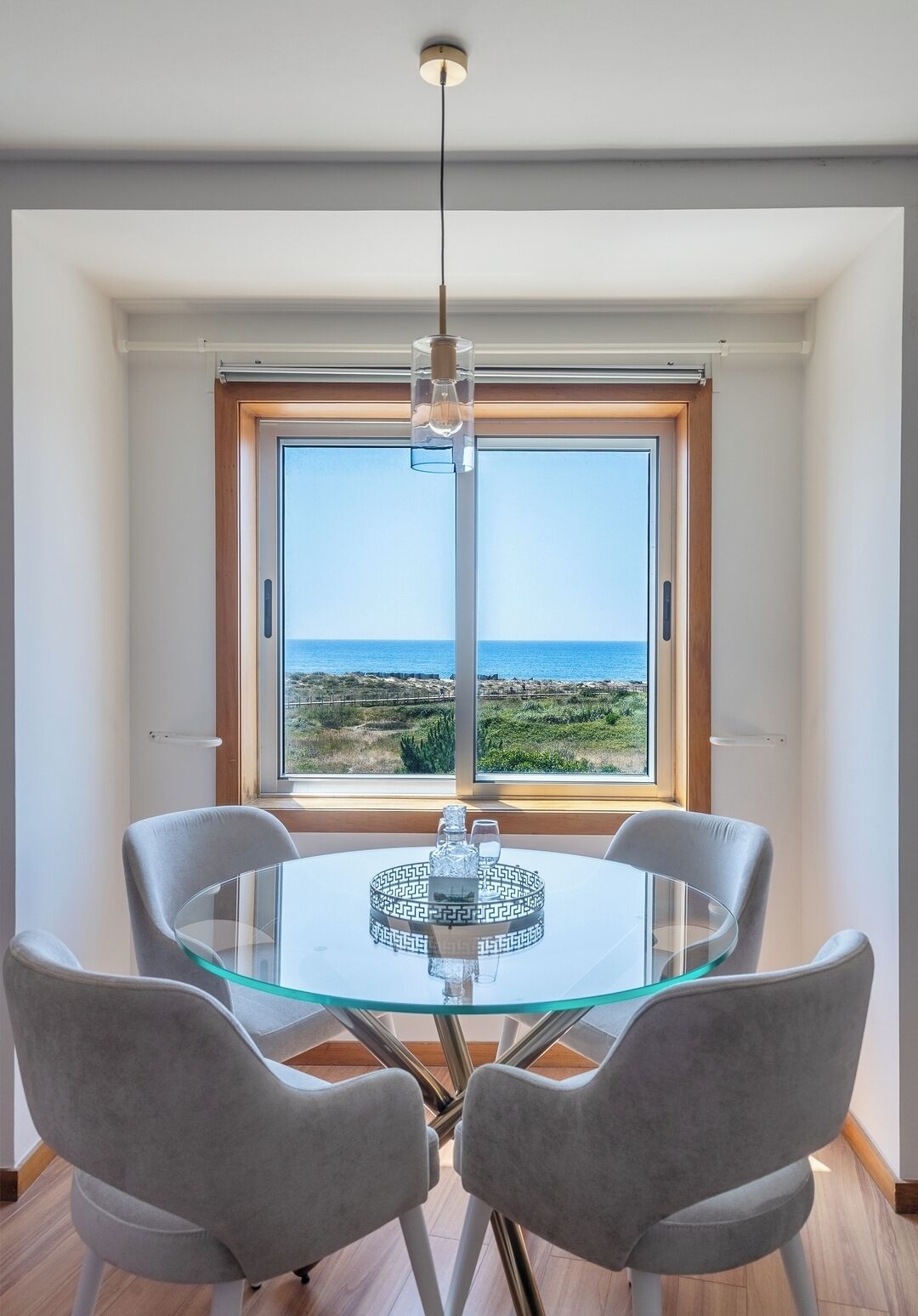 A dining area with a glass table, four grey chairs, a hanging light fixture, and a view of the sea through a large window.