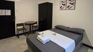 Un letto singolo con lenzuolo bianco, un tavolino con una sedia, un armadio nero e due quadri appesi al muro.