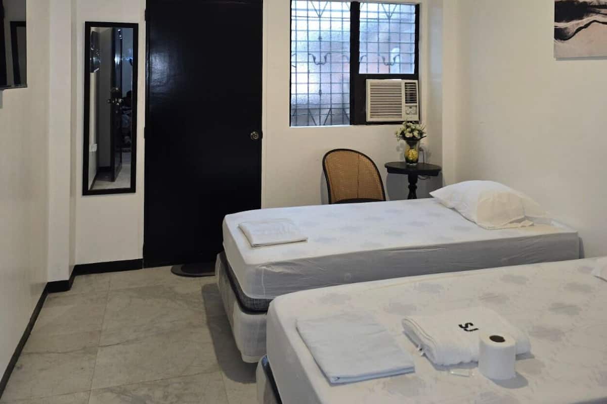 Ein Hotelzimmer mit zwei Betten, einer Tür, einem Fenster mit Klimaanlage, einem Sessel und einem kleinen Tisch mit einer Vase.