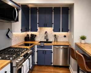 Une cuisine de taille réduite avec des meubles de couleur bleu foncé, un lave-vaisselle en acier inoxydable, une cuisiniÚre à gaz et un four à micro-ondes.