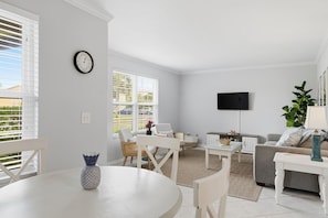 Ein modernes Wohnzimmer mit Esstisch, Sofas, einem Fernseher und einem großen Fenster mit Jalousien.