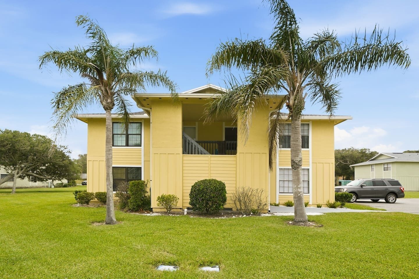 A two-story building with a flat roof, a balcony, and a small front yard with palm trees and shrubs.